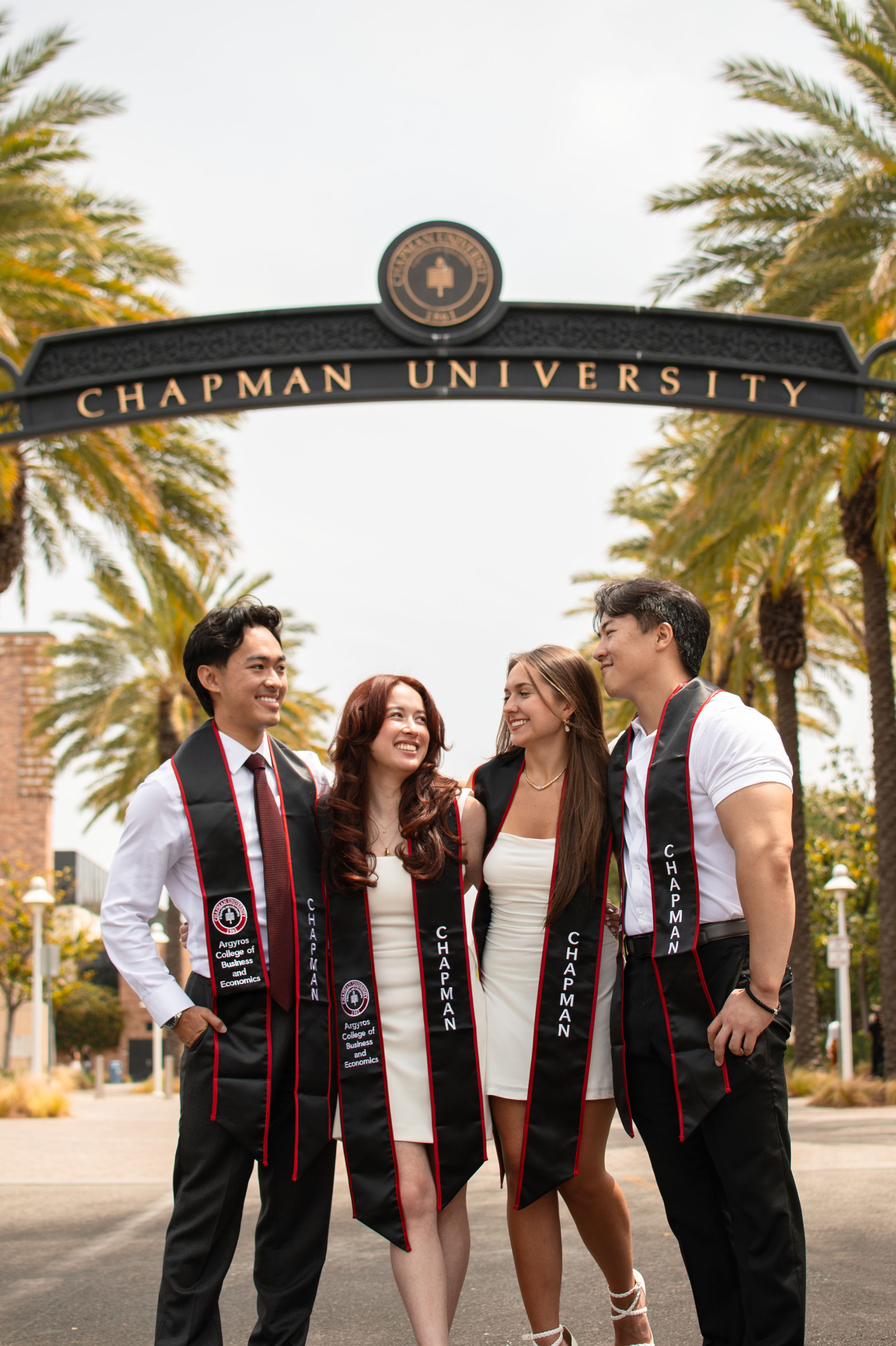 Four Chapman University graduates in white outfits with stoles under campus arch.