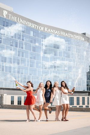 Five women celebrate graduation popping champagne bottles in front of The University of Kansas Health System building.