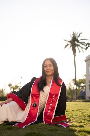 Female graduate sits on green lawn wearing black graduation robe with red Chapman University stole and palm tree behind.