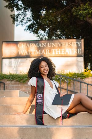 Chapman University graduate smiles on steps near Waltmar Theatre sign wearing white dress and College of Performing Arts stole.
