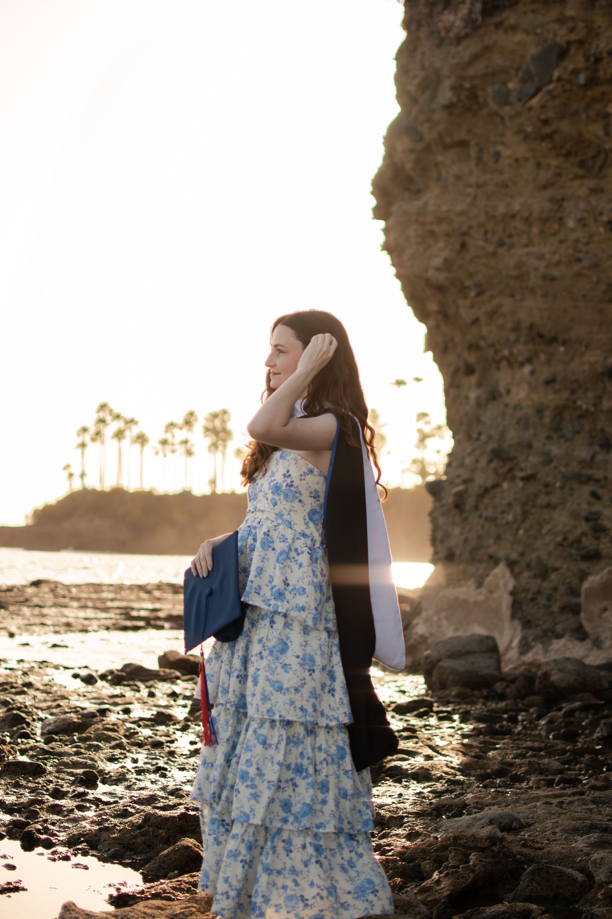 Graduate in floral dress holds cap and gown at sunset beach near rocky cliffs with palm trees in background.