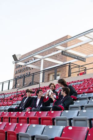 Five male graduates in black suits sit together laughing in red stadium bleachers at Chapman University athletics complex.