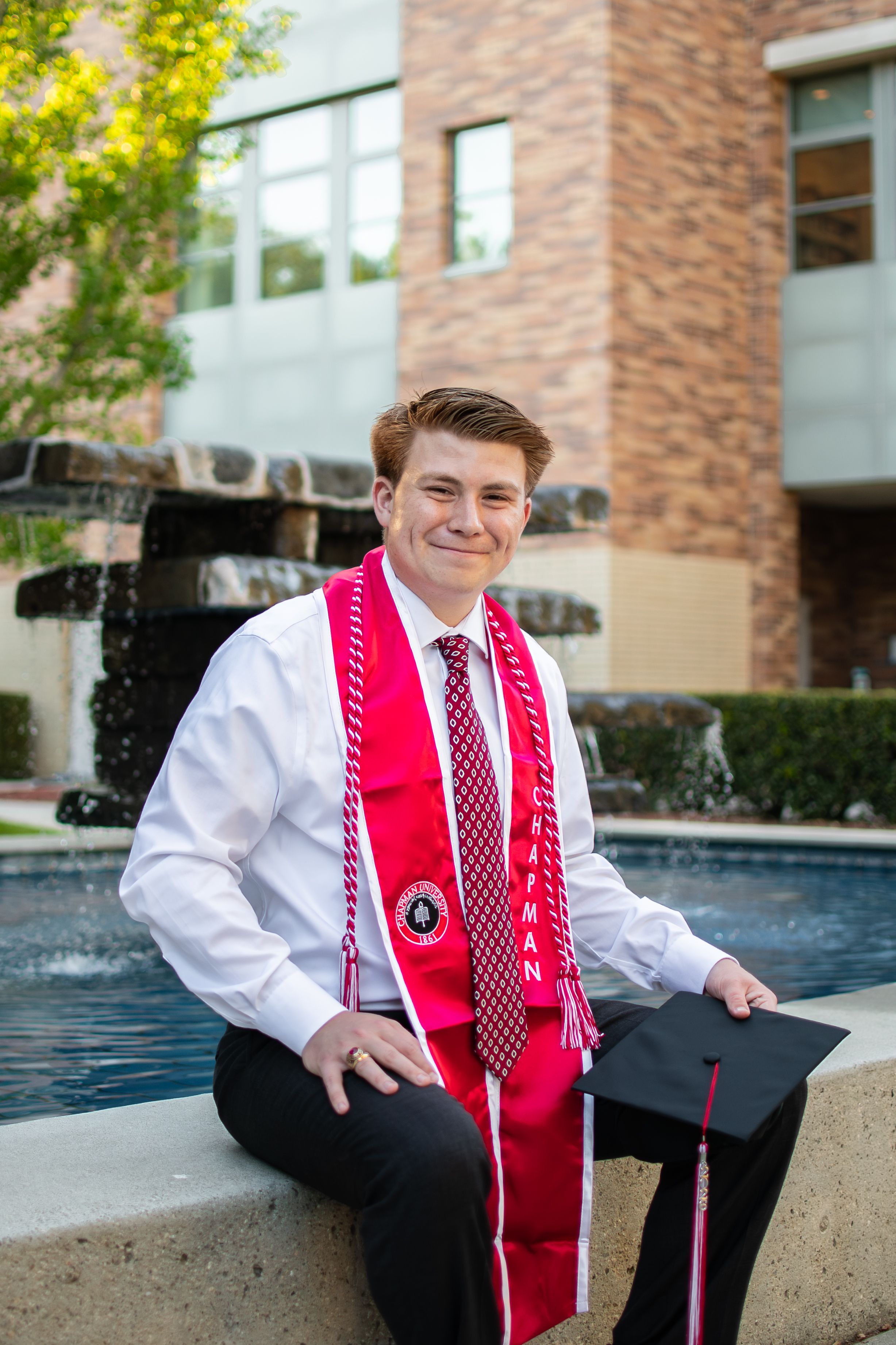 Male Chapman University graduate sits by fountain wearing white shirt, red tie, and red Chapman graduation stole holding cap.