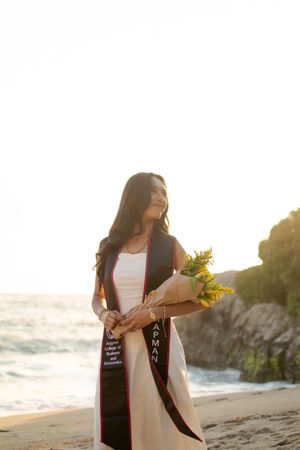 Female Chapman University graduate stands on beach at sunset holding yellow sunflowers wearing white dress and graduation stole.