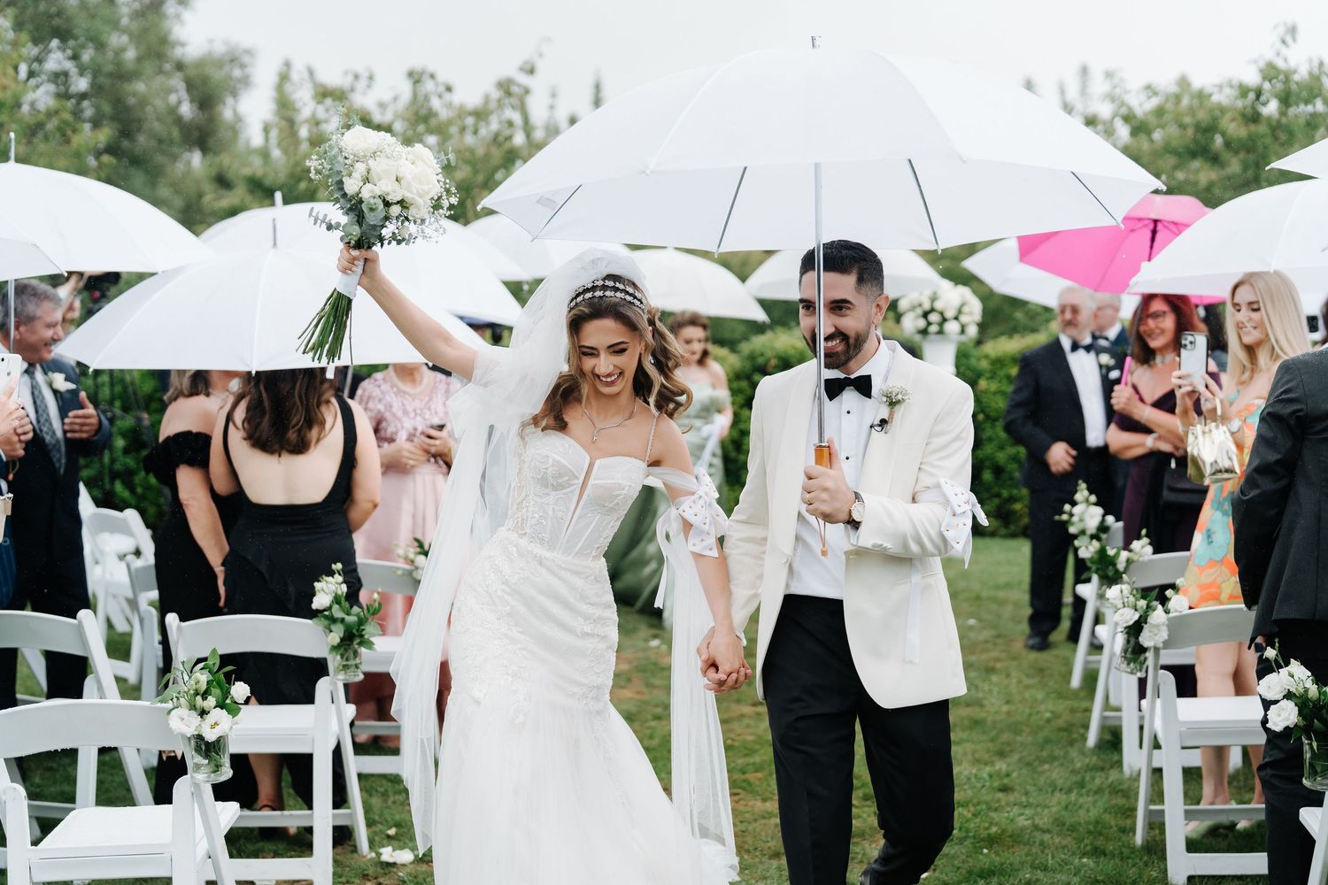 Bride and groom walk down outdoor aisle holding hands under white umbrellas surrounded by guests at elegant wedding.