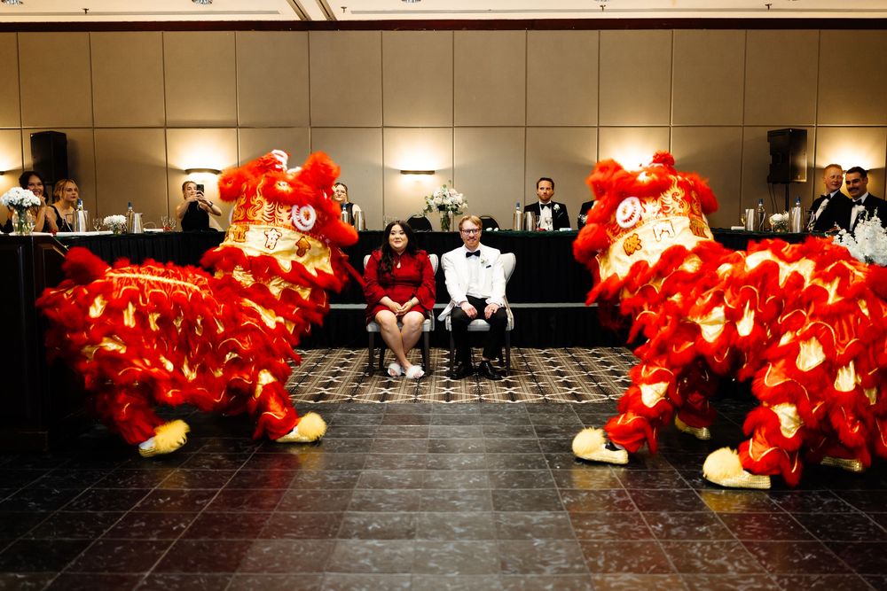 Two people sit between vibrant red Chinese lion dance costumes during a festive indoor celebration.