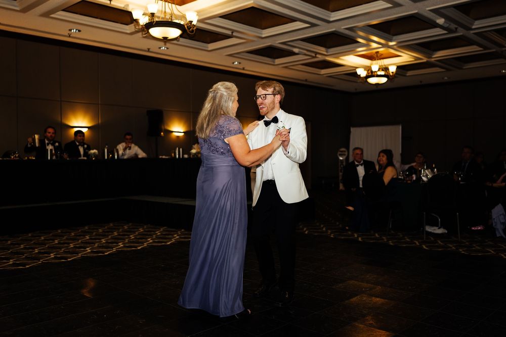 Groom in white tuxedo shares mother-son dance at elegant wedding reception in dimly lit ballroom.