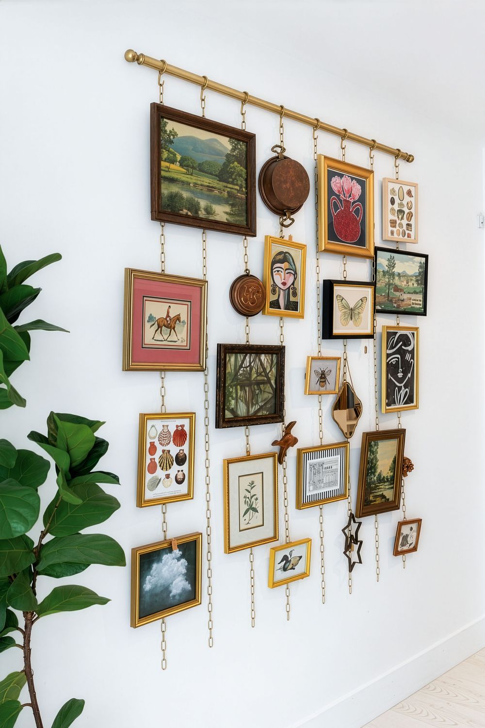 A gallery wall display featuring eclectic framed artwork hung from brass chains on a gold rod against a white wall. Swell House La Grange