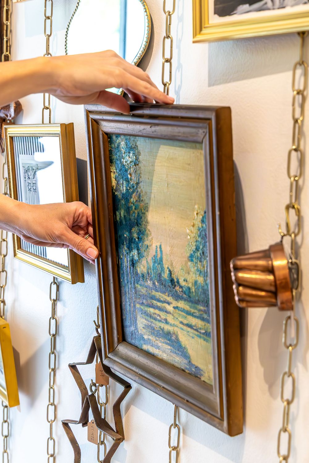 Hands adjusting a vintage framed landscape painting on a wall decorated with gold chains and antique decor. Swell House La Grange