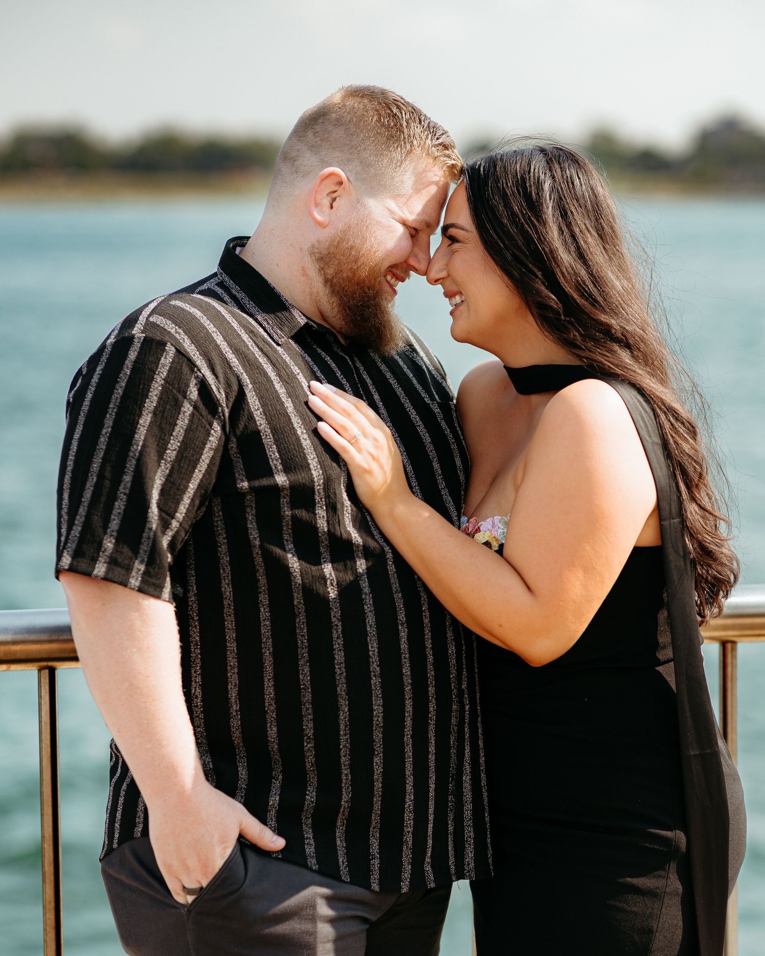 Couple in black outfits sharing a romantic moment by the waterfront, touching foreheads and smiling.
