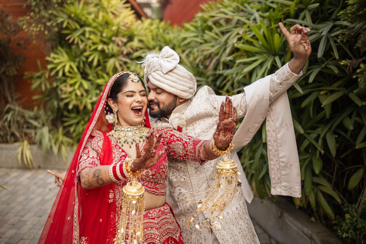 Indian bride and groom in traditional red and white wedding attire dancing joyfully outdoors.