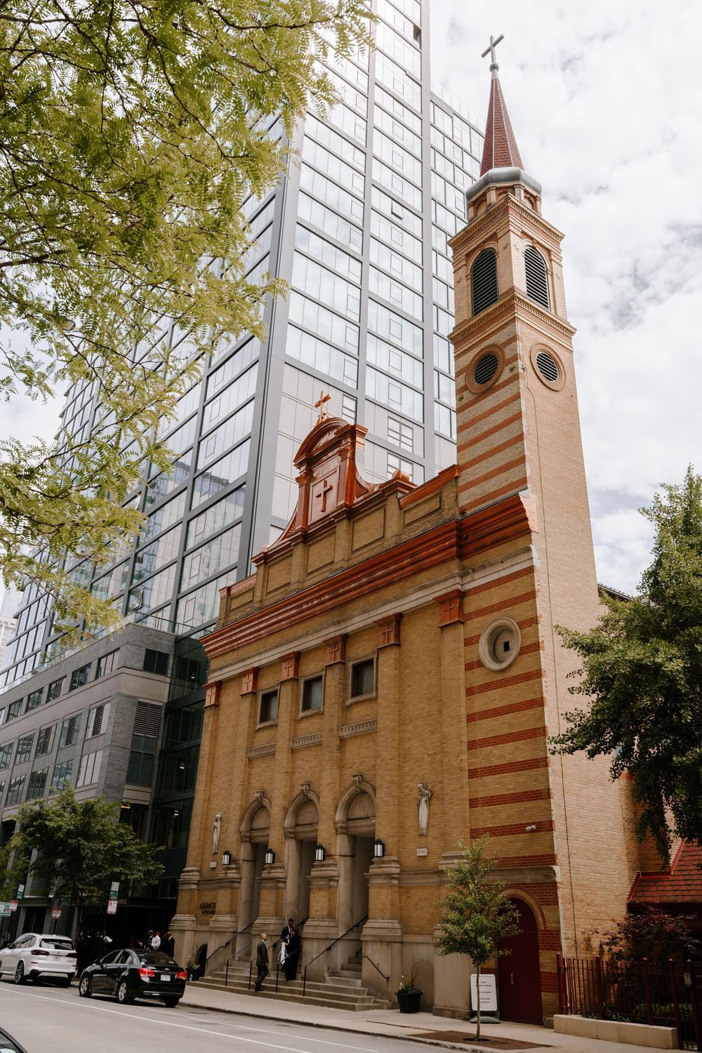 Historic brick church with bell tower and cross stands beside modern glass skyscraper in urban setting.
