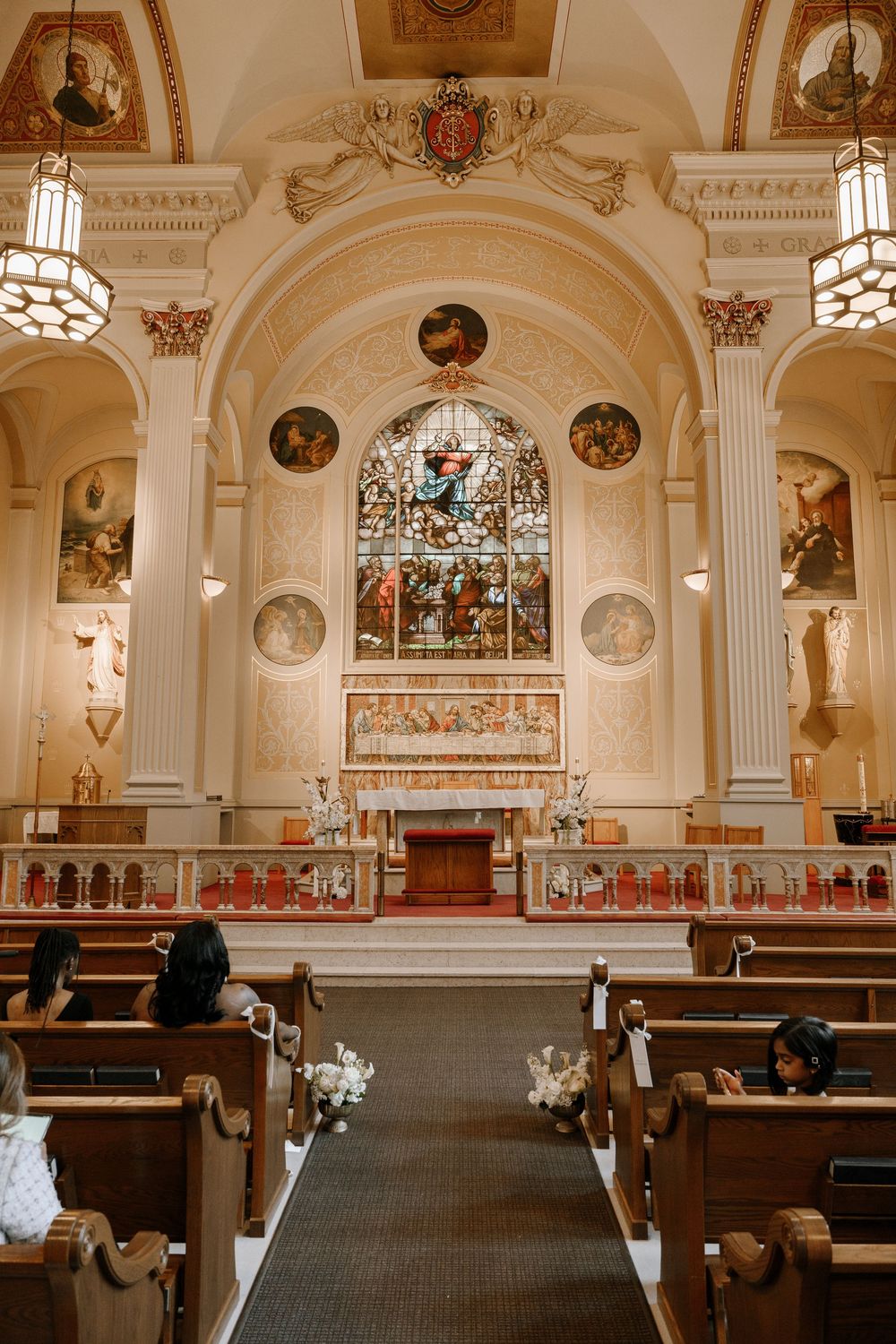 Elegant Catholic church interior with ornate altar, stained glass window, and wooden pews decorated for a wedding ceremony.