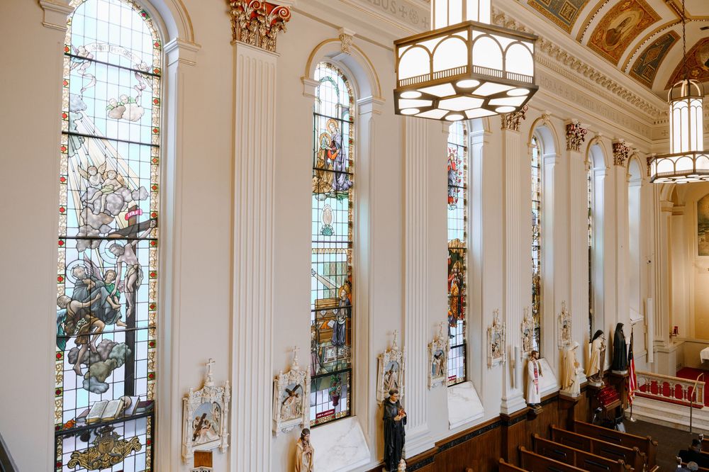 Ornate church interior featuring tall stained glass windows, white columns with gold details, and hanging chandeliers.