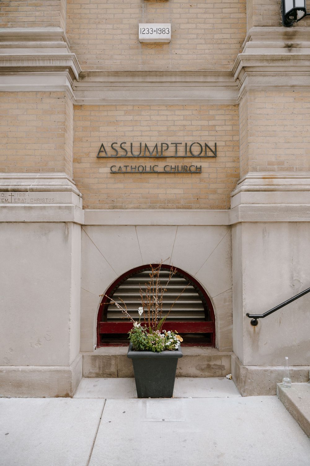Assumption Catholic Church exterior entrance with stone facade, arched window, and a flower planter on the sidewalk.