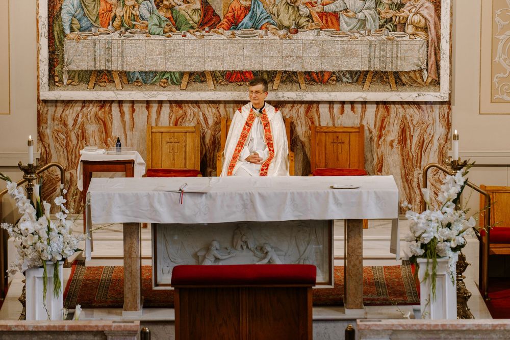 Priest in white and orange vestments stands at ornate altar with floral arrangements beneath a Last Supper tapestry.
