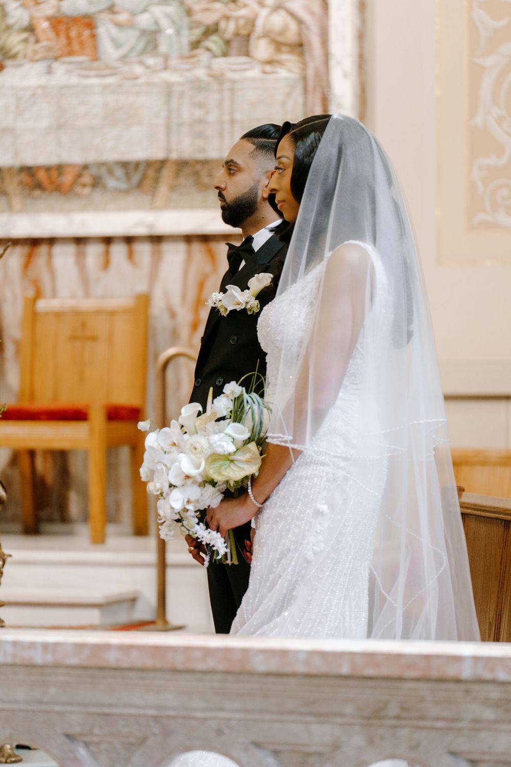Bride in beaded gown holding white floral bouquet stands beside groom in black suit at church altar.