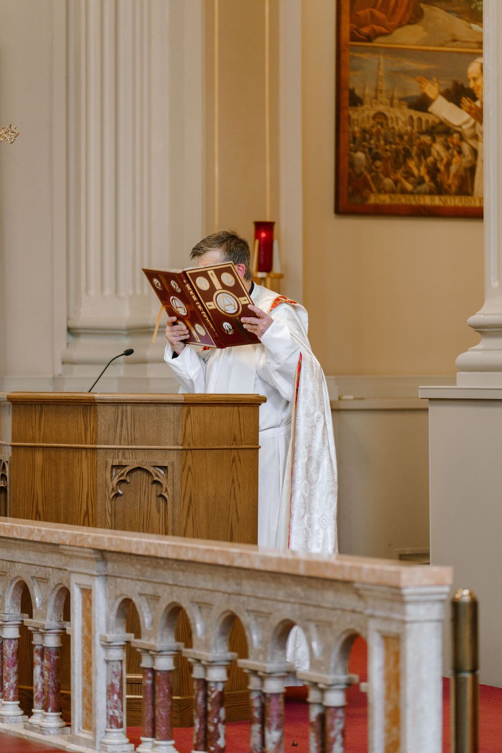 Catholic priest elevating the Roman Missal during Mass at an ornate church altar with marble railings.