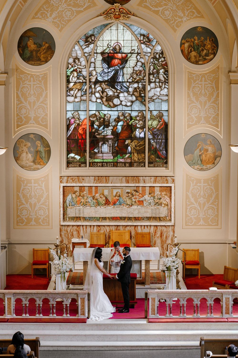 Bride and groom exchange vows at ornate church altar with stunning stained glass window and Last Supper artwork.