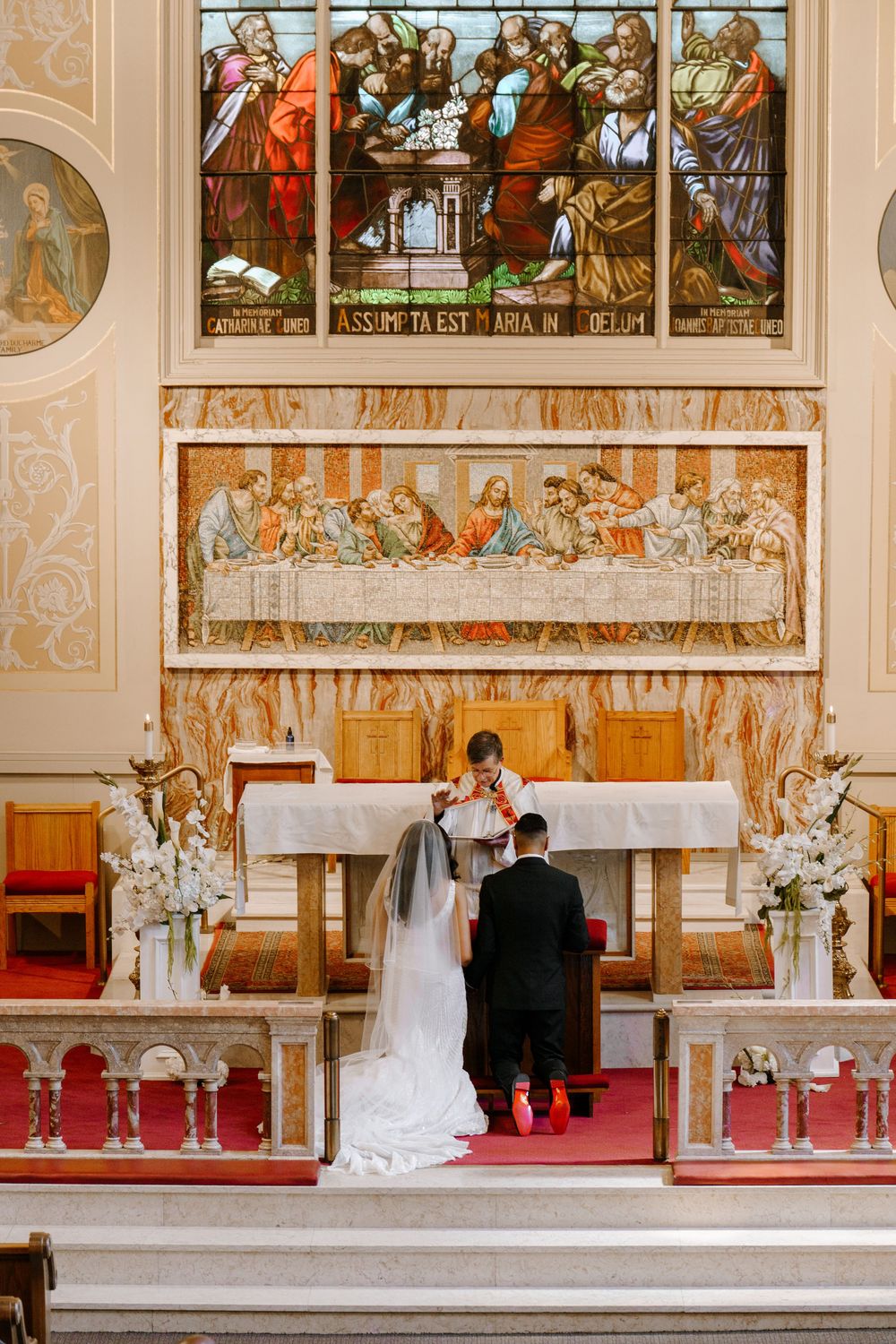 Bride and groom at ornate church altar with Last Supper mosaic and stained glass window during wedding ceremony.