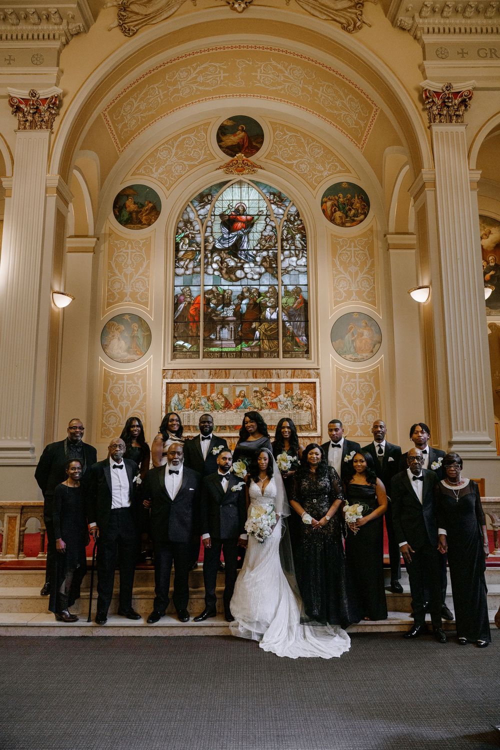 Wedding party poses in elegant church with stained glass window, bride in white gown surrounded by guests in black formal wear.