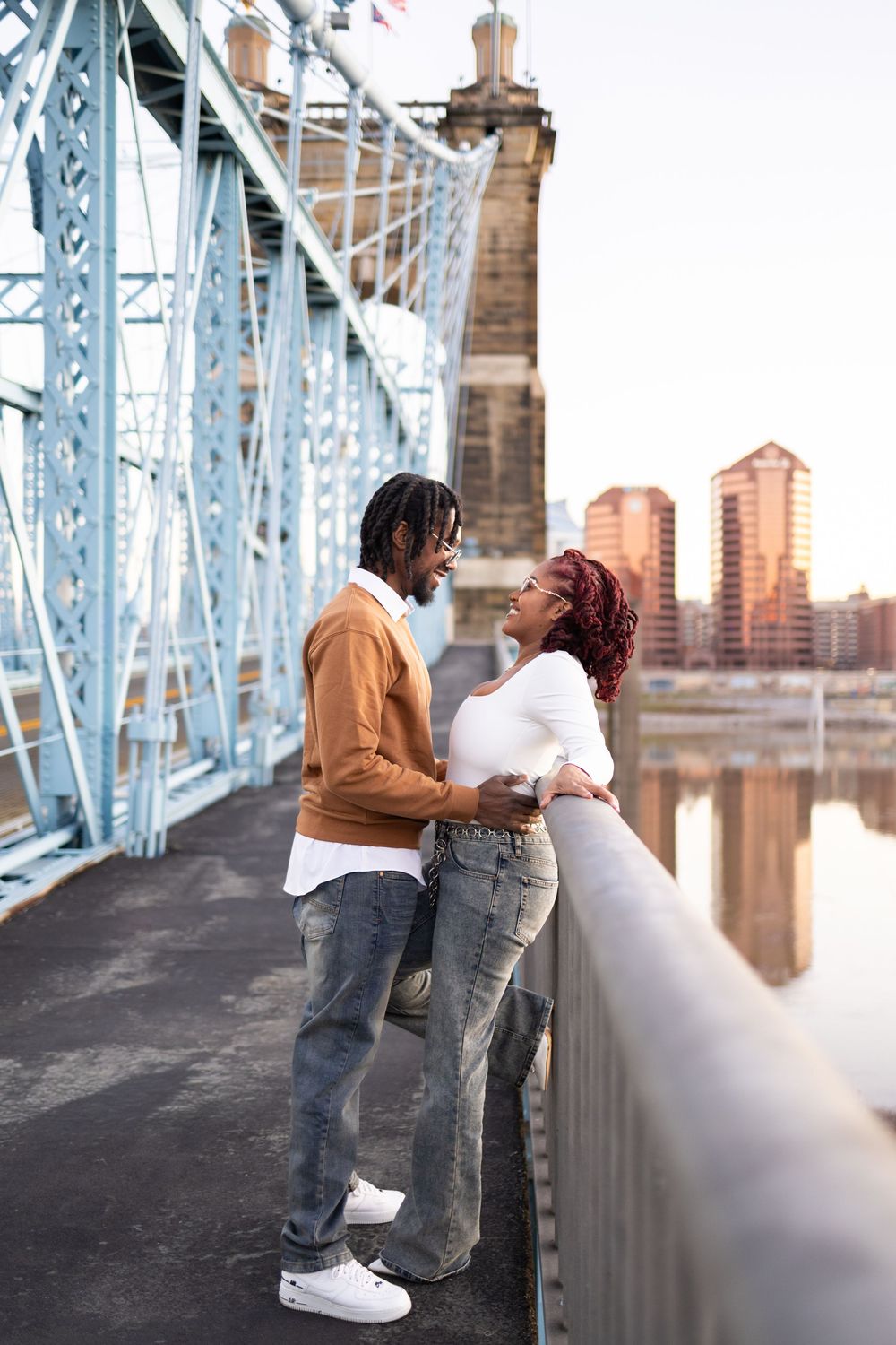 A couple shares a romantic moment in Smale Park in Cincinnati on a bridge with city skyline and river views in the background.