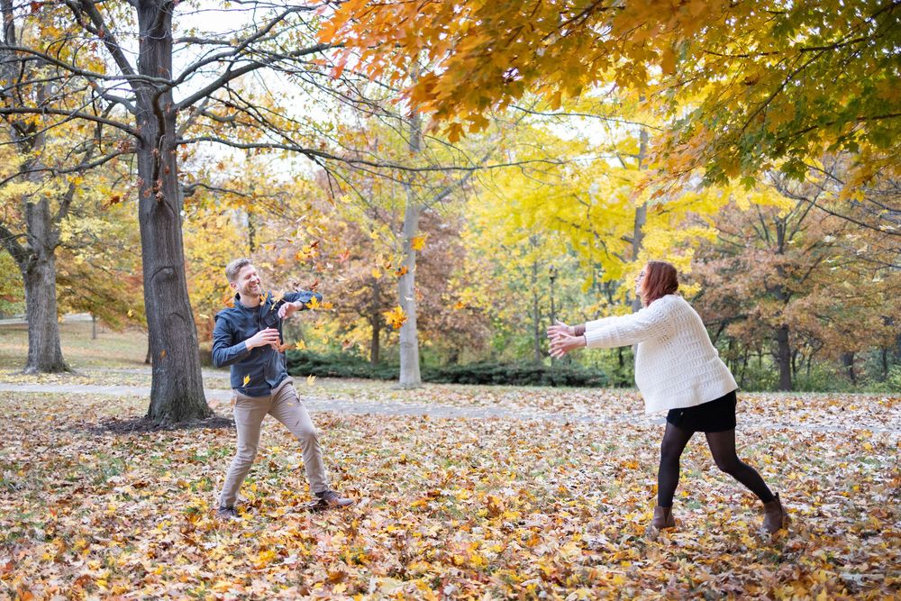 Two people playfully tossing autumn leaves in a colorful fall park surrounded by yellow and orange foliage.