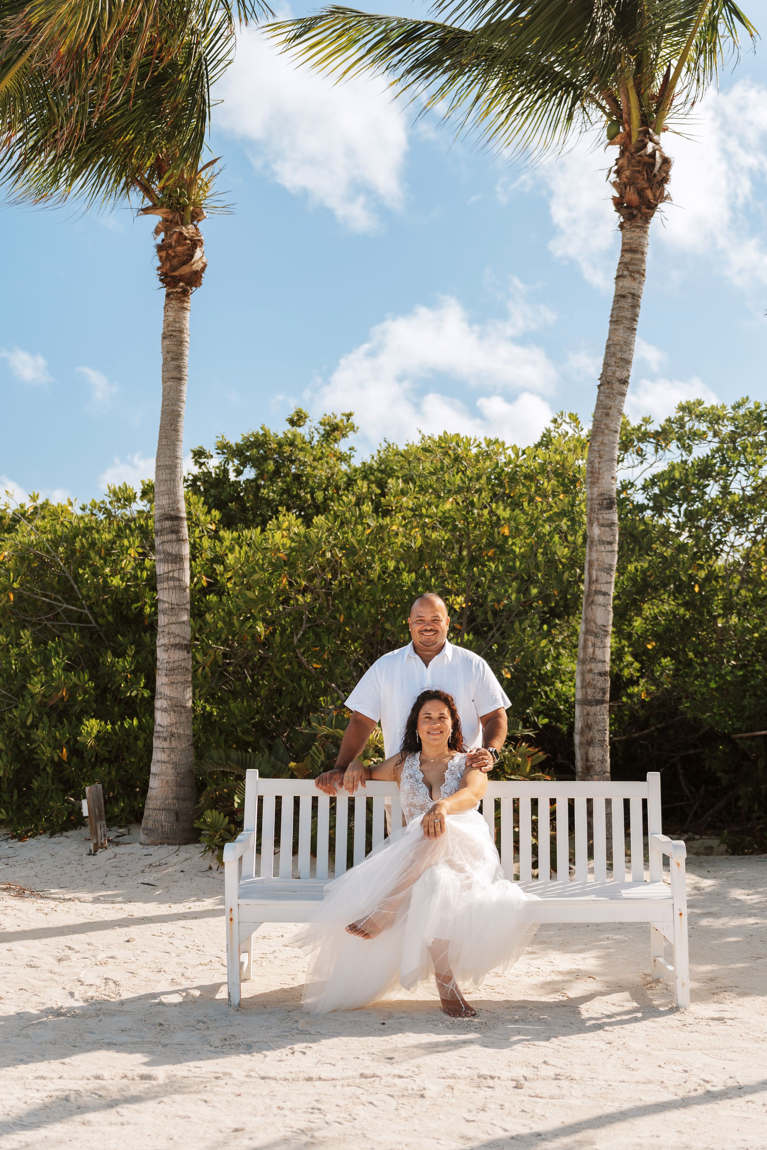 Couple posing on a white bench between palm trees on a sandy beach, woman in white dress, man in white shirt.