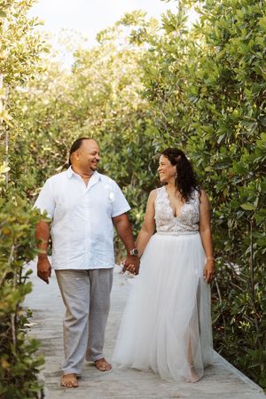 Bride and groom walk hand-in-hand along a tropical boardwalk path, surrounded by lush green foliage.