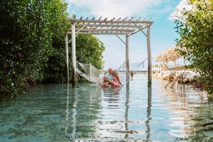 Person relaxing in a hammock over crystal-clear turquoise water surrounded by lush tropical trees and a white pergola.
