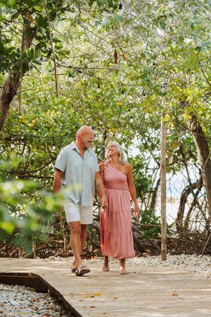 A couple walks hand-in-hand on a wooden boardwalk surrounded by lush tropical trees.