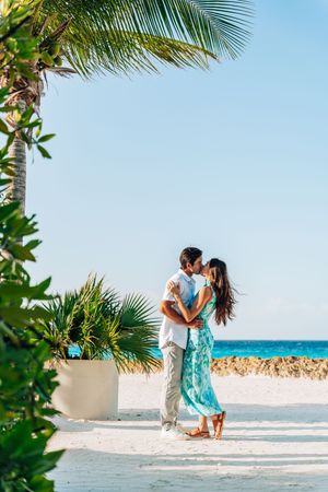 Romantic couple kissing on a tropical beach with palm trees and turquoise ocean in the background.