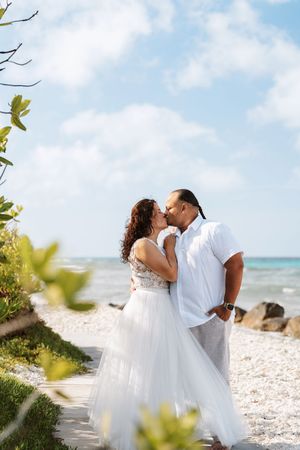 Couple sharing a romantic kiss on a sandy beach path, bride in white tulle gown, groom in white linen, ocean backdrop.