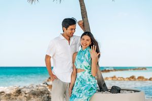 Smiling couple in tropical beach setting, man in white shirt and woman in teal floral dress waving near palm tree.
