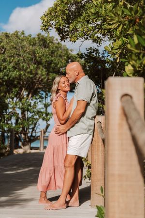 Couple embracing on a tropical wooden deck, woman in pink dress, man in sage shirt, surrounded by lush greenery.
