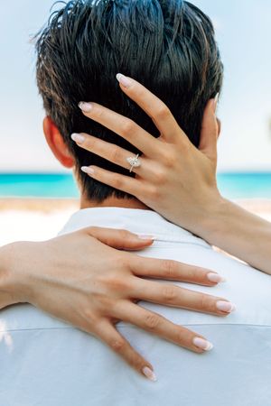 Woman with engagement ring embracing man from behind on a tropical beach, showcasing diamond ring on her finger.