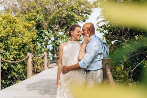 Couple sharing a romantic moment on a tropical boardwalk, surrounded by lush greenery, dressed in elegant white and blue.
