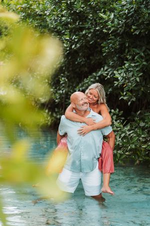 A couple embraces in turquoise water surrounded by lush greenery, man giving woman a piggyback ride.