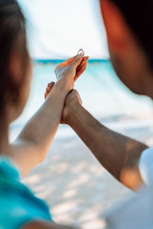 Couple holding hands on a beach while man presents a diamond engagement ring during a romantic proposal.