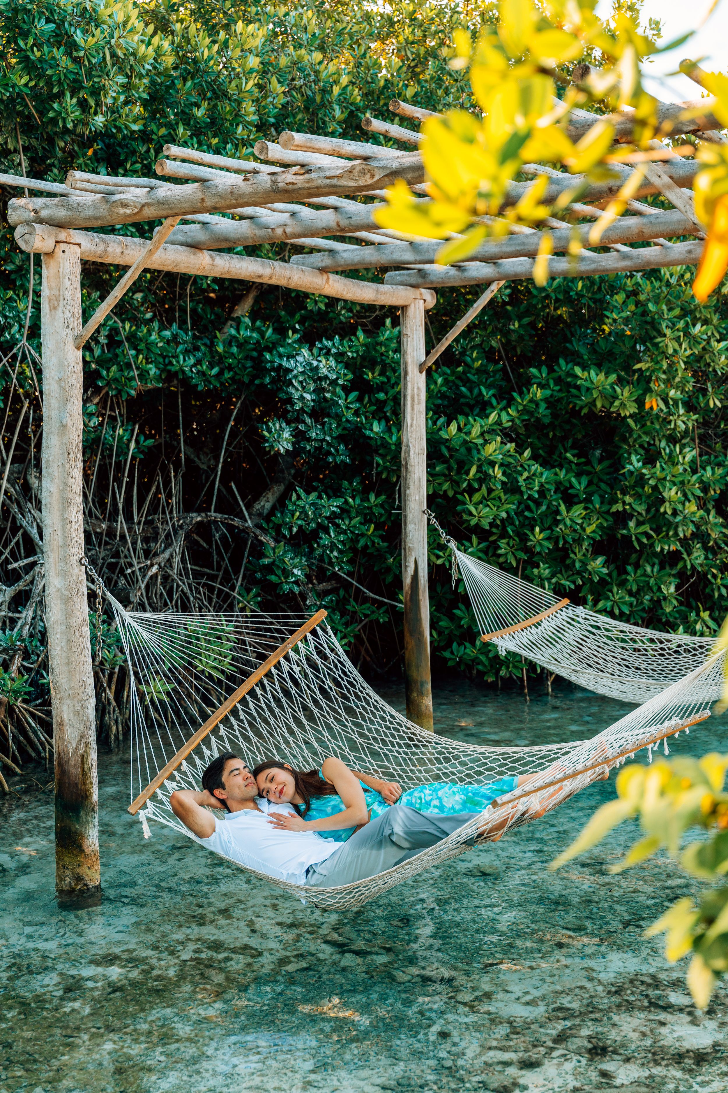Couple relaxing together in a rope hammock under a wooden pergola surrounded by lush tropical greenery.