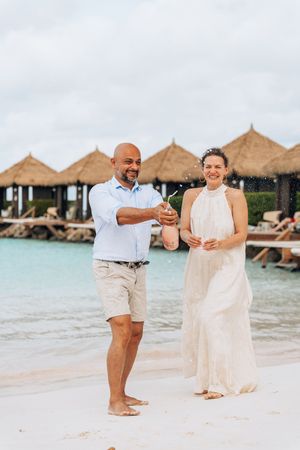 Couple celebrating on a tropical beach, popping champagne near overwater bungalows with turquoise water.