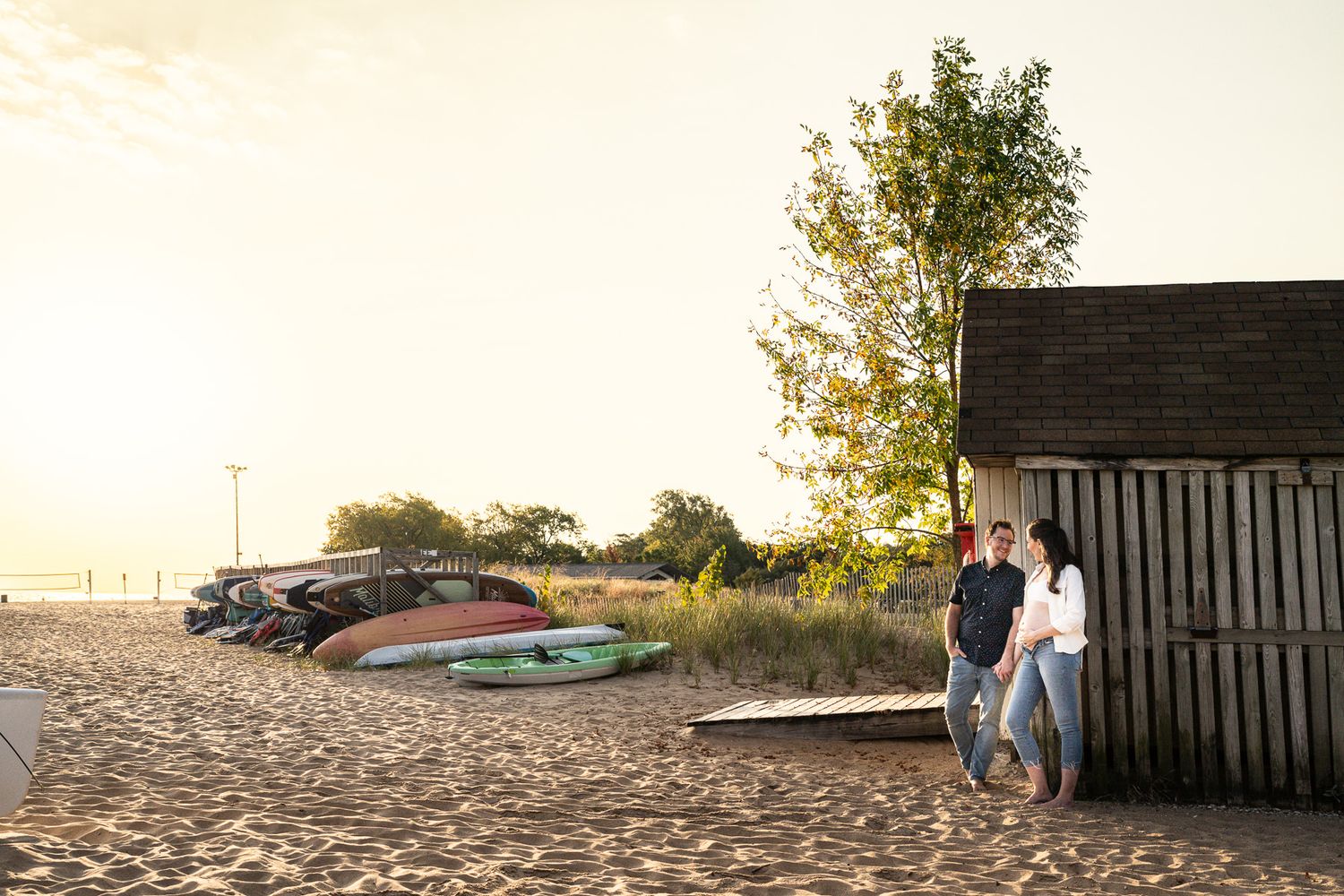 Couple leaning against rustic beach shed at golden hour with colorful kayaks on sandy shore.