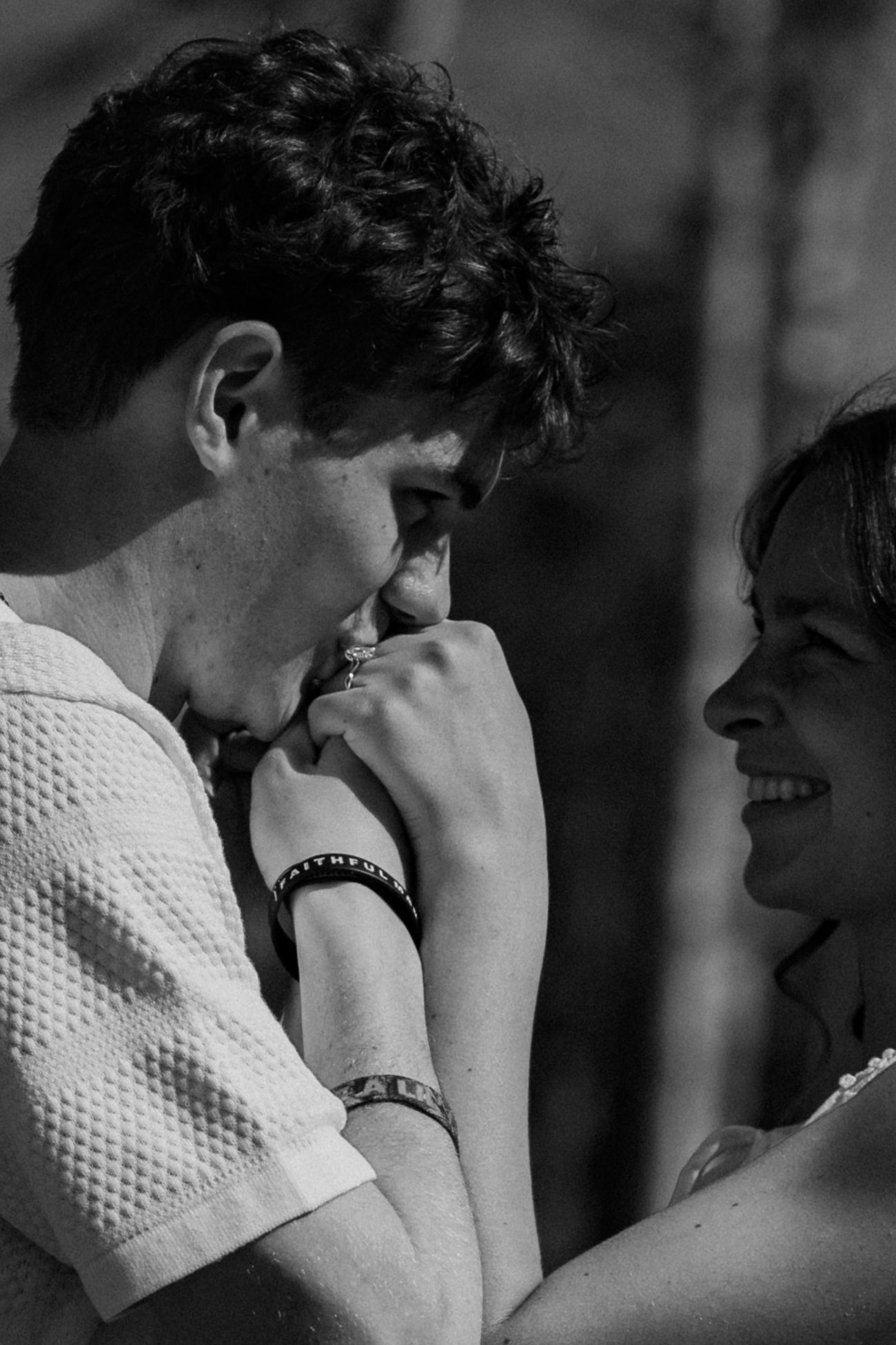 Black and white photo of a young couple sharing a tender moment, smiling and holding hands outdoors.