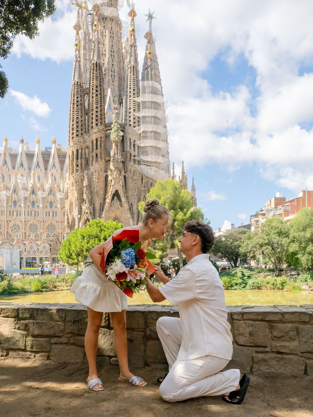 Romantic marriage proposal in front of Sagrada Familia, Barcelona, man kneeling with bouquet.