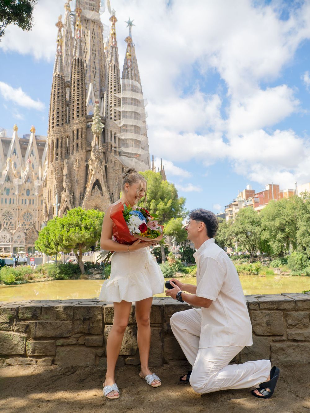 Man proposing to woman with flowers in front of Sagrada Familia, Barcelona, on a sunny day.