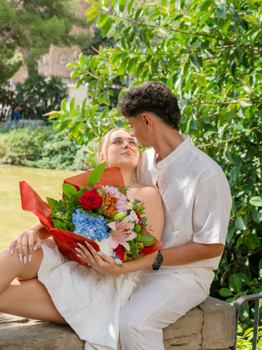 A couple sits on a stone bench in a garden, sharing a romantic moment with a colorful bouquet of flowers.