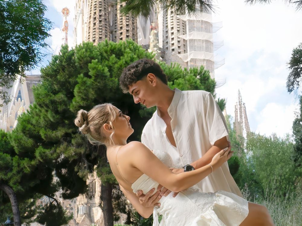 Romantic couple dancing near Sagrada Familia in Barcelona, dressed in white outfits among lush green trees.