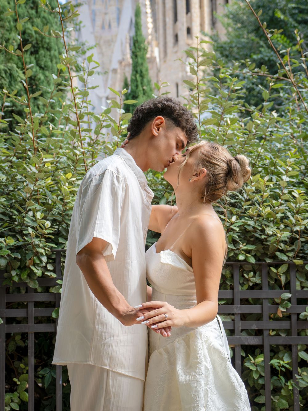Couple in white attire sharing a romantic moment near lush greenery with architectural details in background.