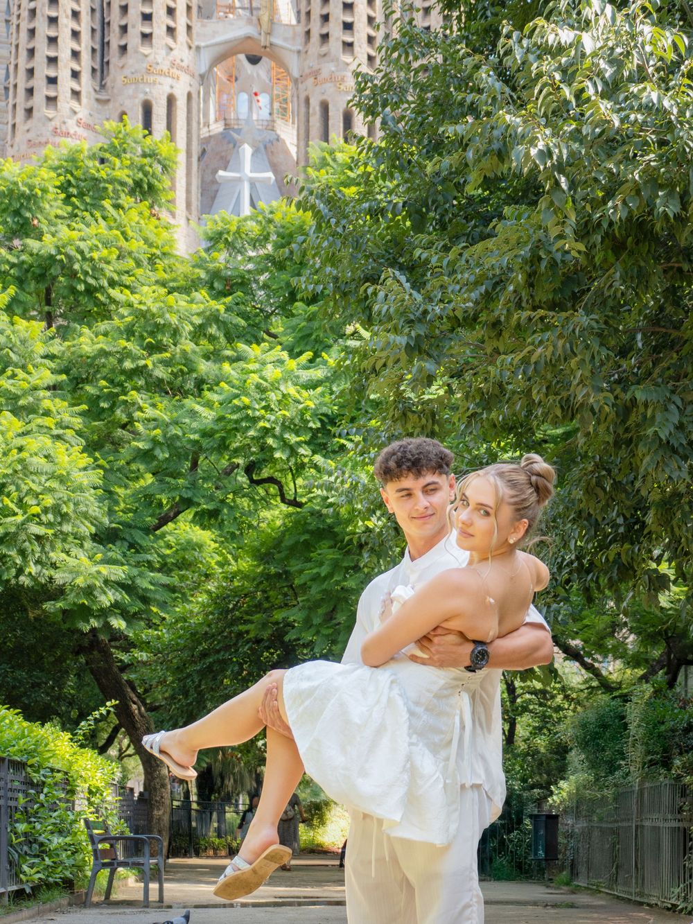 Couple posing romantically in front of Sagrada Familia, Barcelona, man lifting woman in white dress outdoors.
