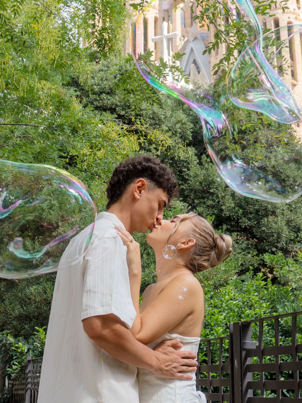 Couple kissing surrounded by giant soap bubbles with lush green trees and architecture in the background.